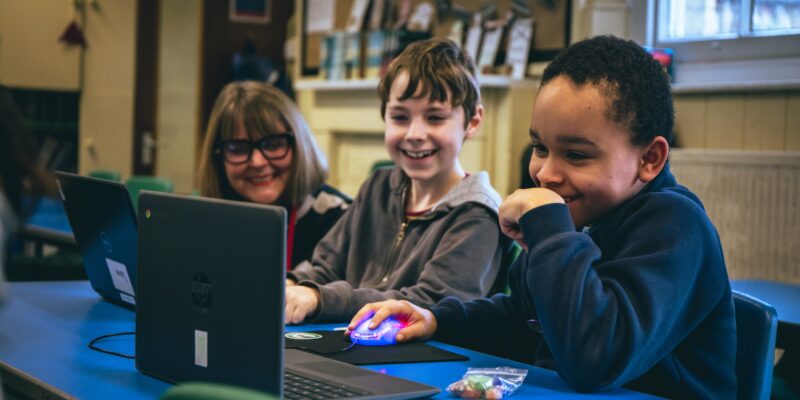 Two young people smiling whilst working on their laptop with an adult mentor by their side.